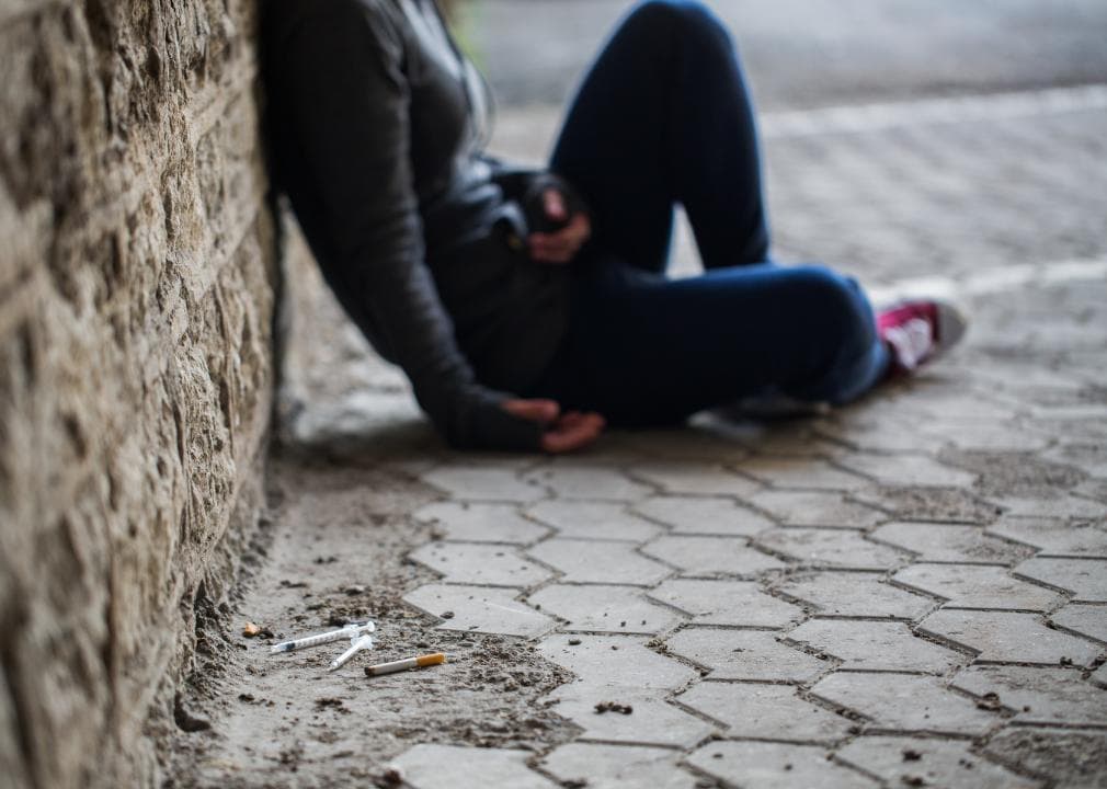 person sitting in the sidewalk next to syringes