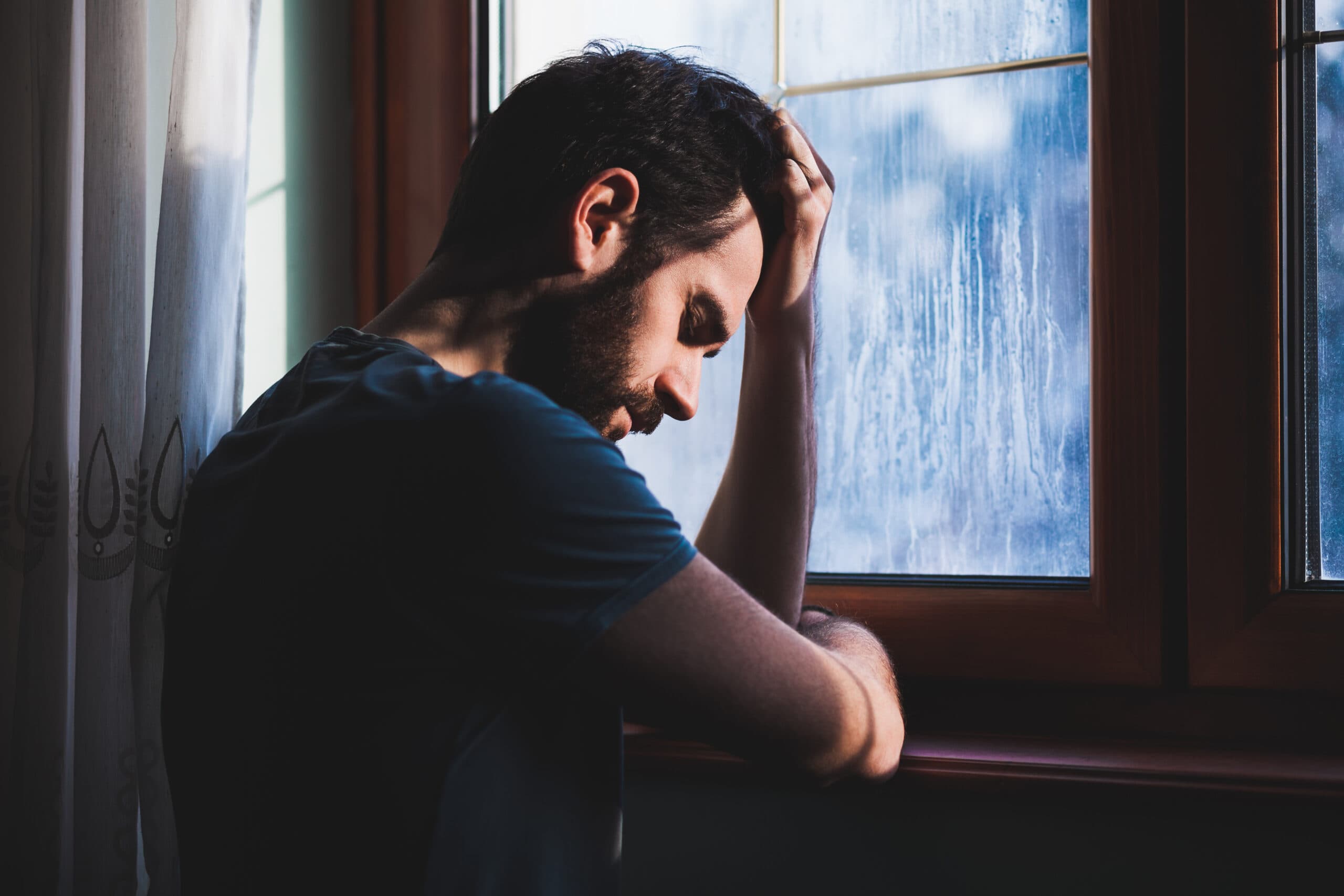 man with head in hand standing at window in pain