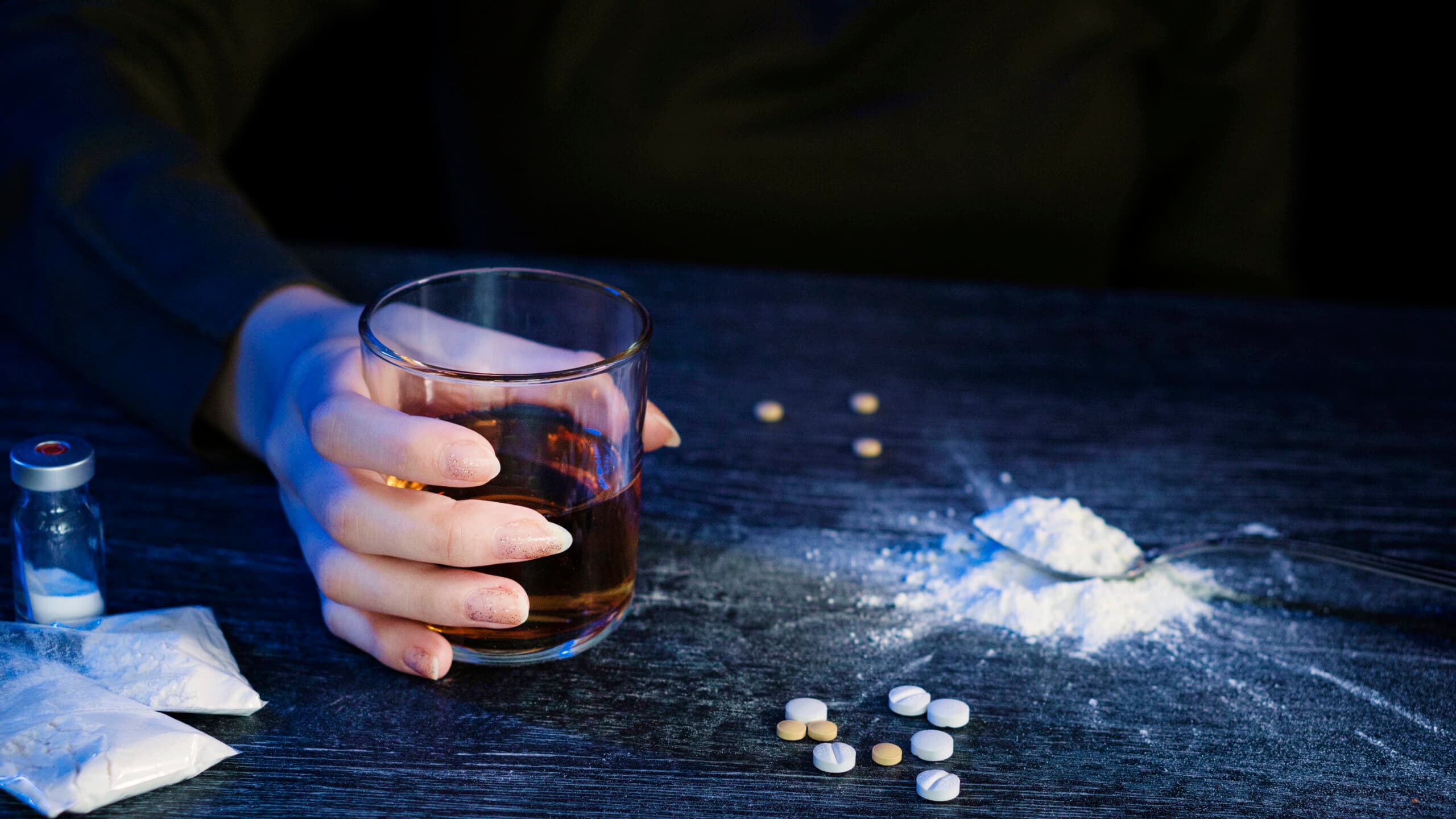 woman holding glass of liquor with powder drugs and pills on table