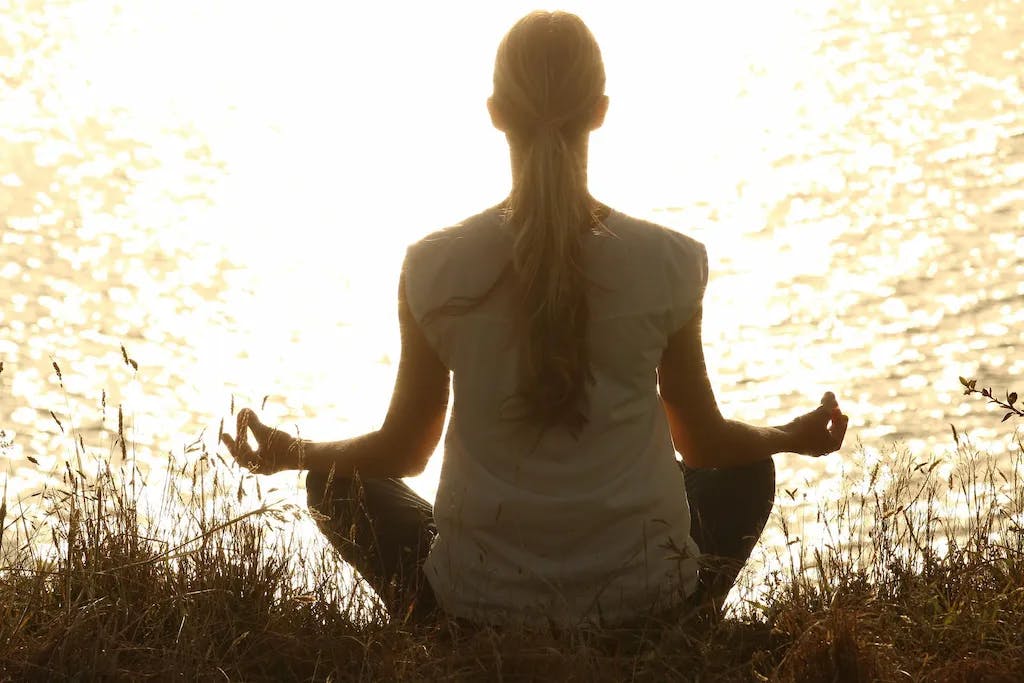Woman sits in lotus position at edge of lake during sunrise.