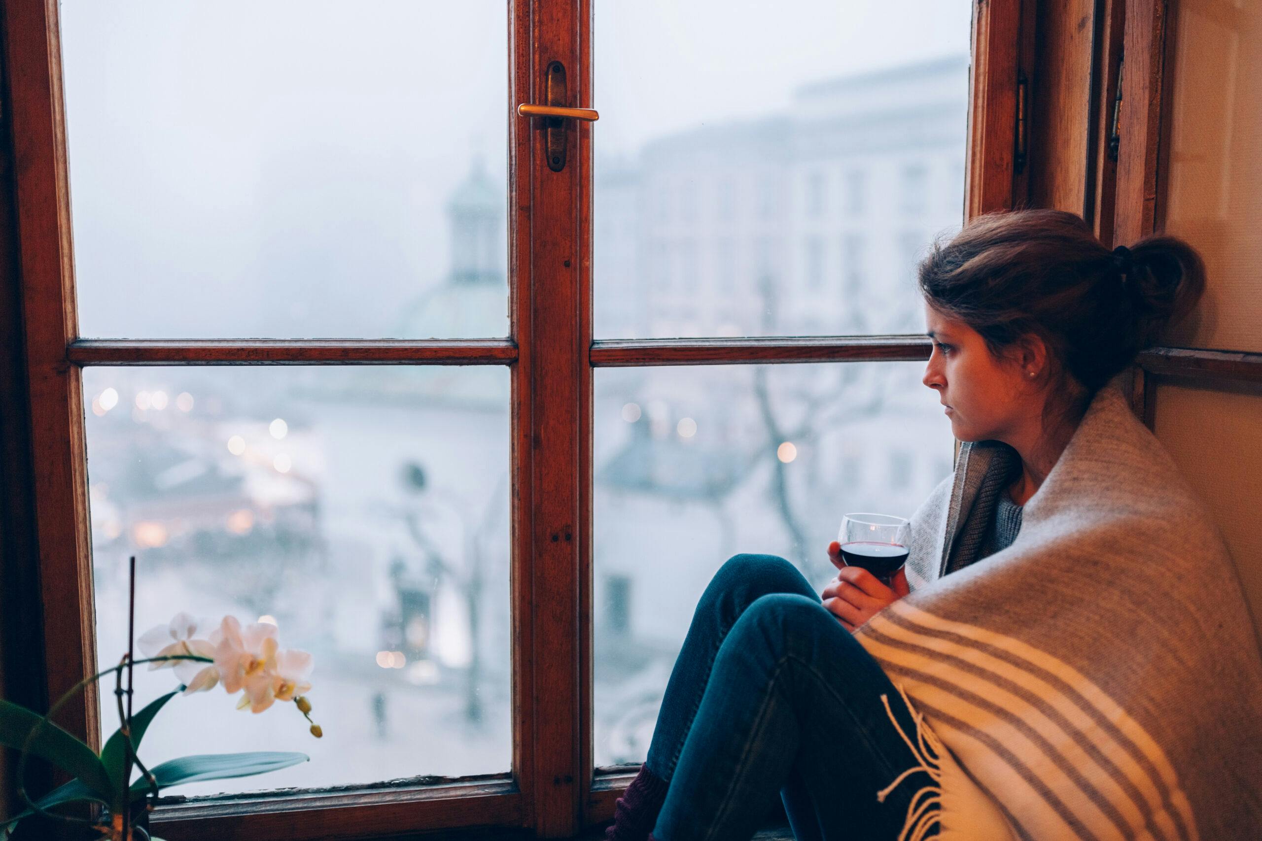 depressed woman looking out window with glass of wine