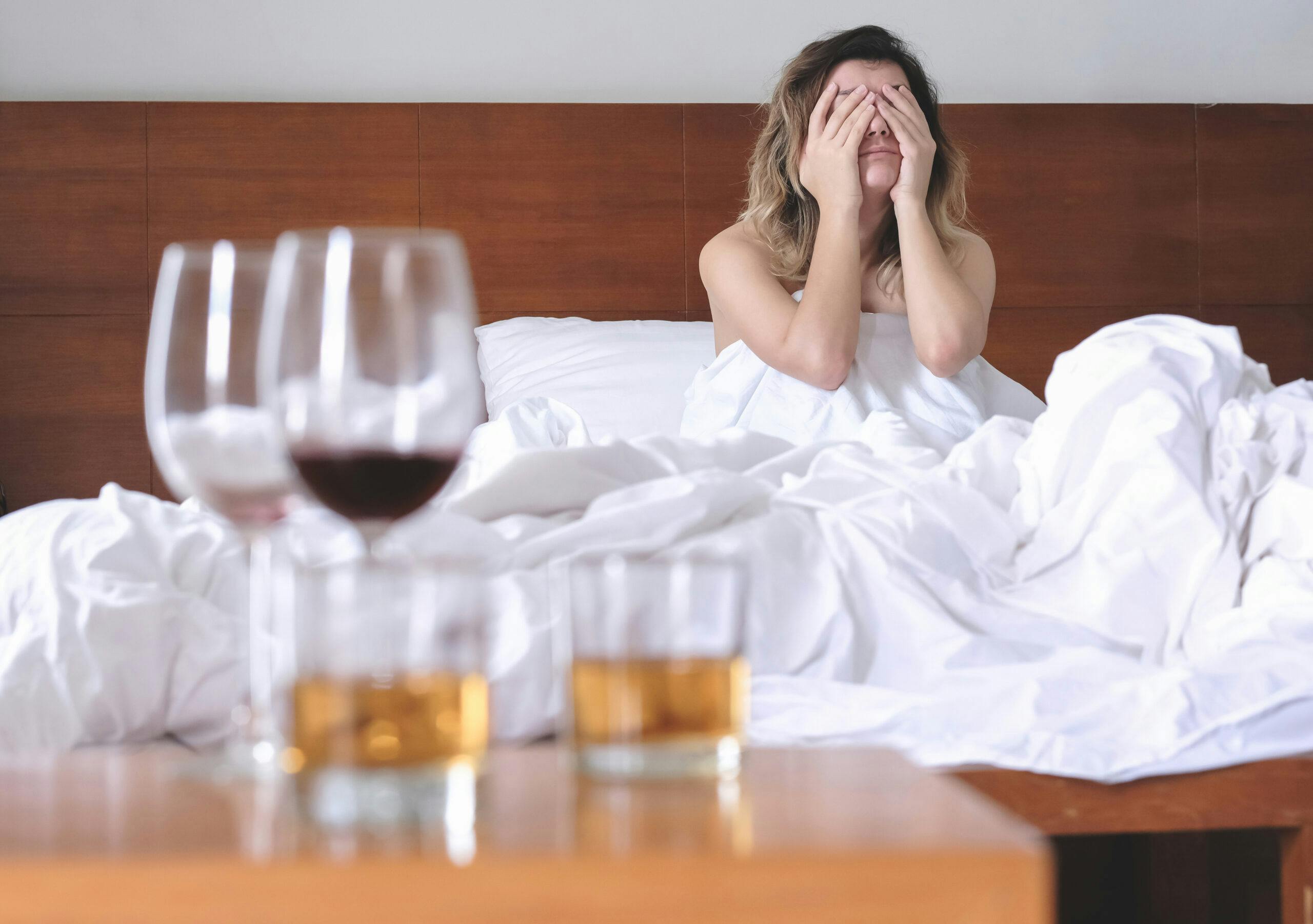 young woman waking up in hotel bed with liquor and wine glasses hungover