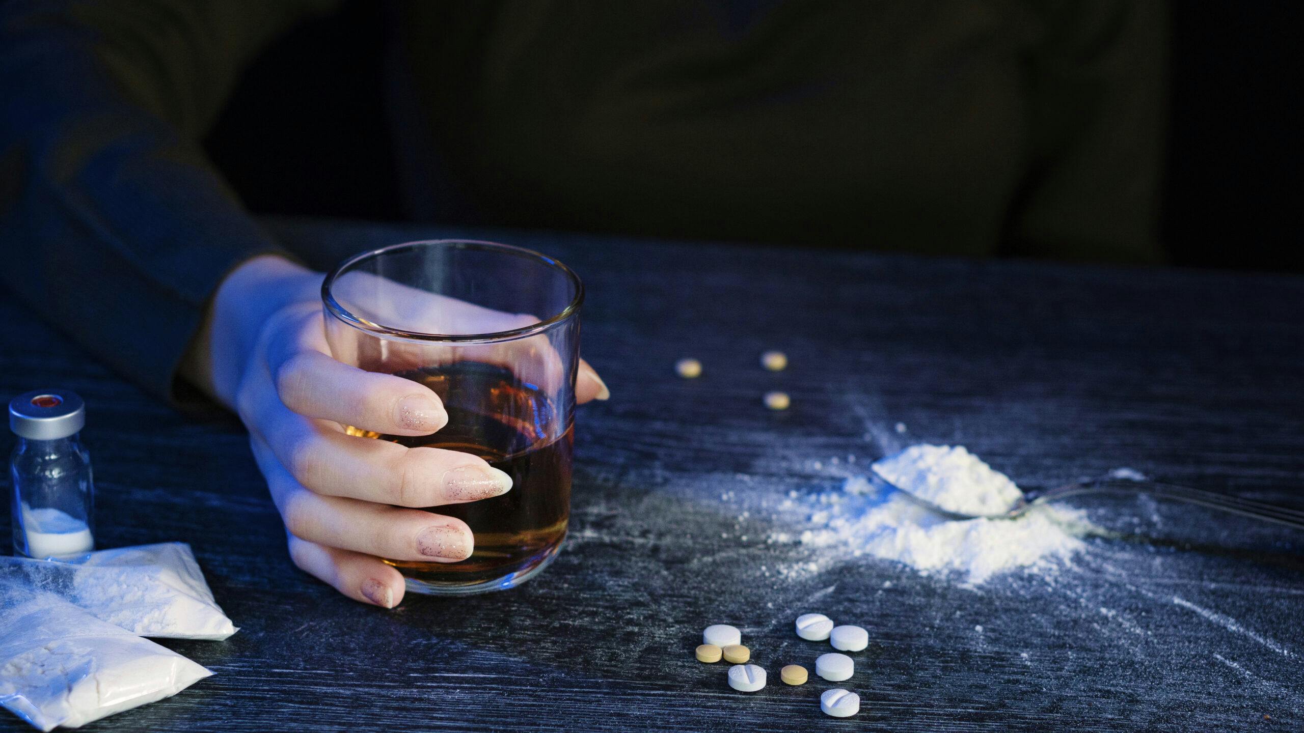 woman holding glass of liquor with powder drugs and pills on table